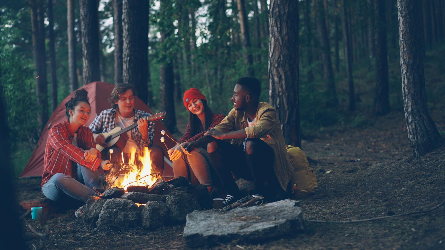 Group of friends camping, playing guitar, and roasting marshmallows by a campfire.