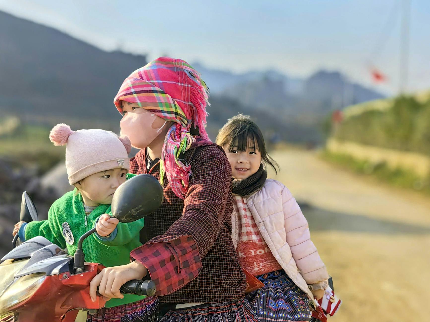 A family of three riding a scooter down a rural road in Vietnam, showcasing cultural attire.