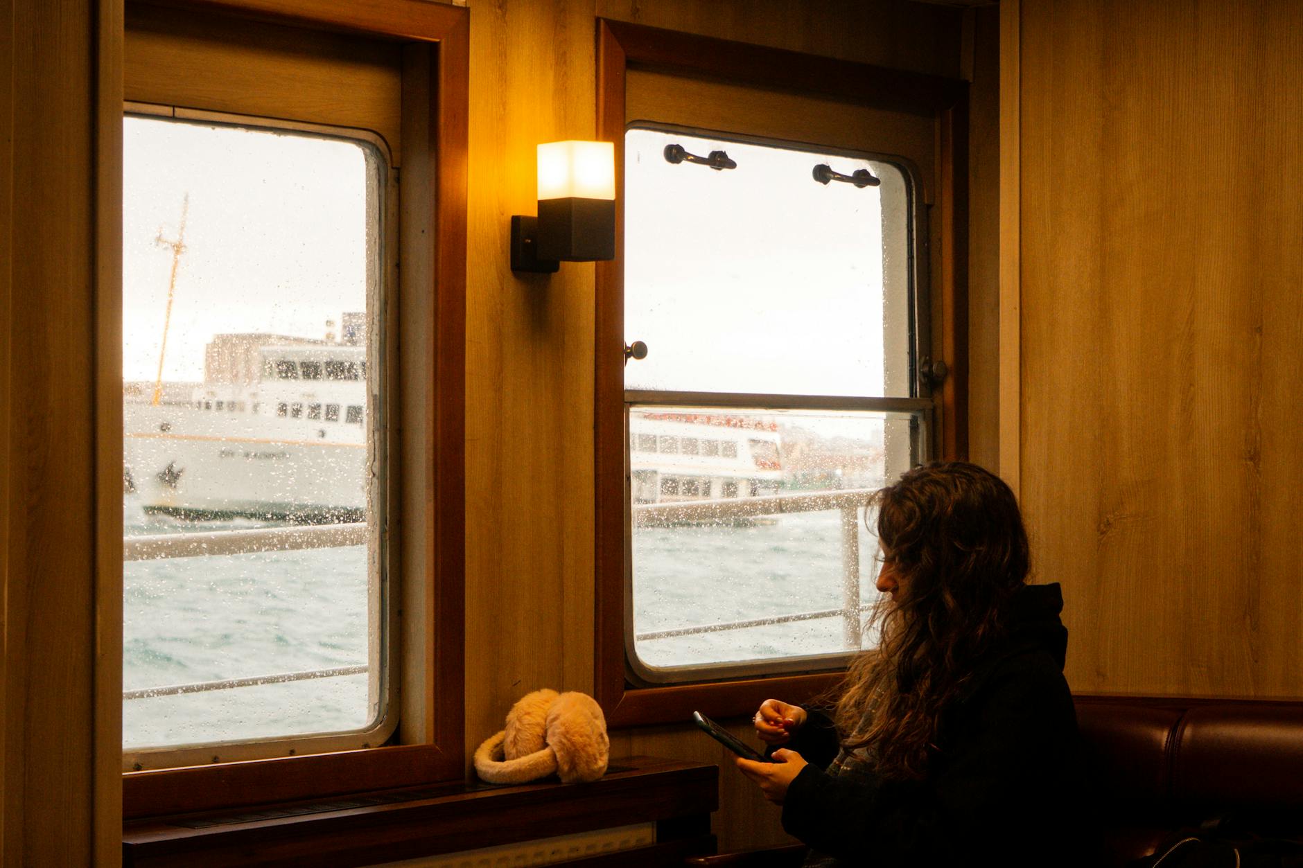 Woman sitting inside a ferry with ocean view and boats visible through window.