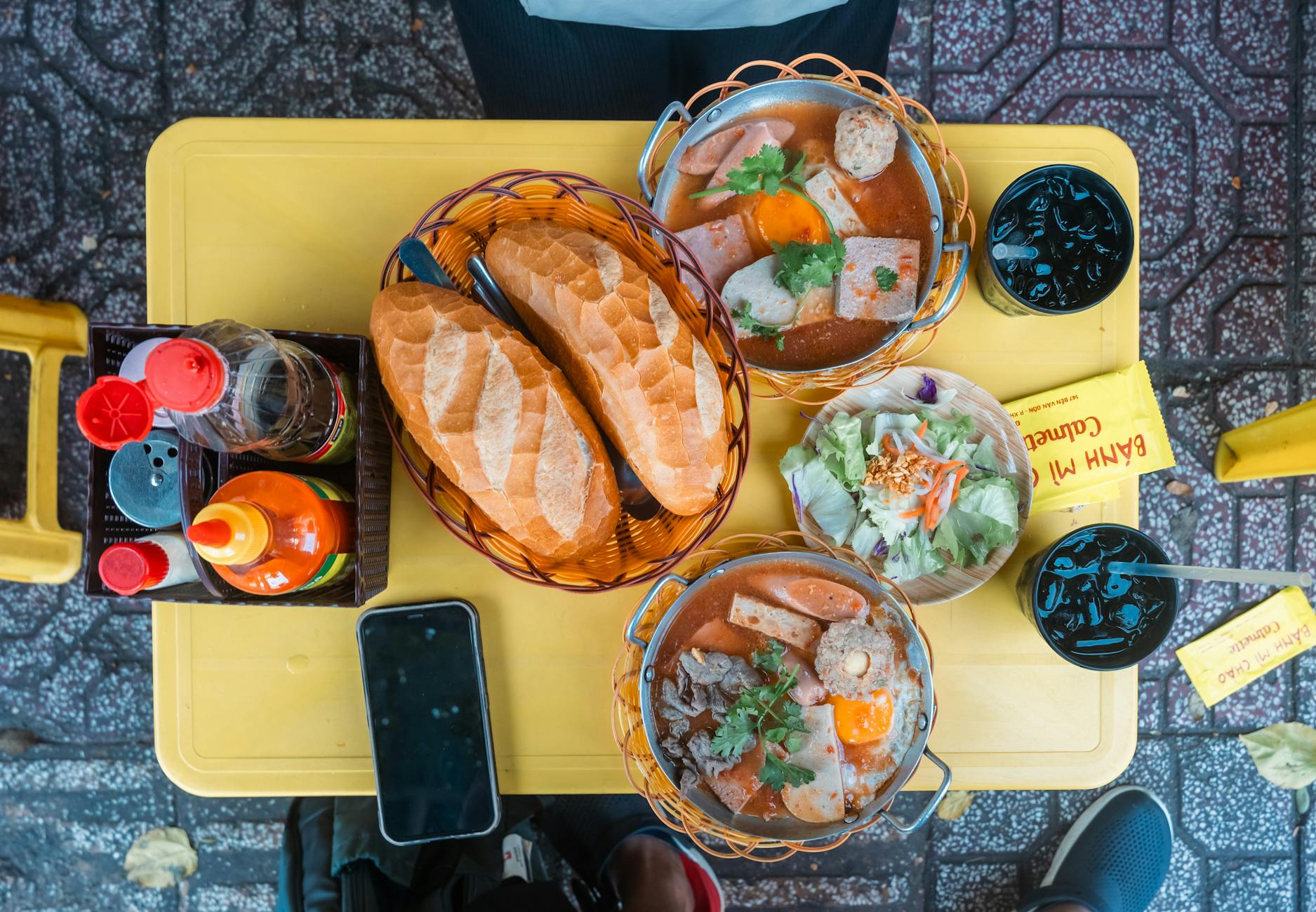 Colorful display of Vietnamese street food with bread, soup, and drinks on a yellow table.