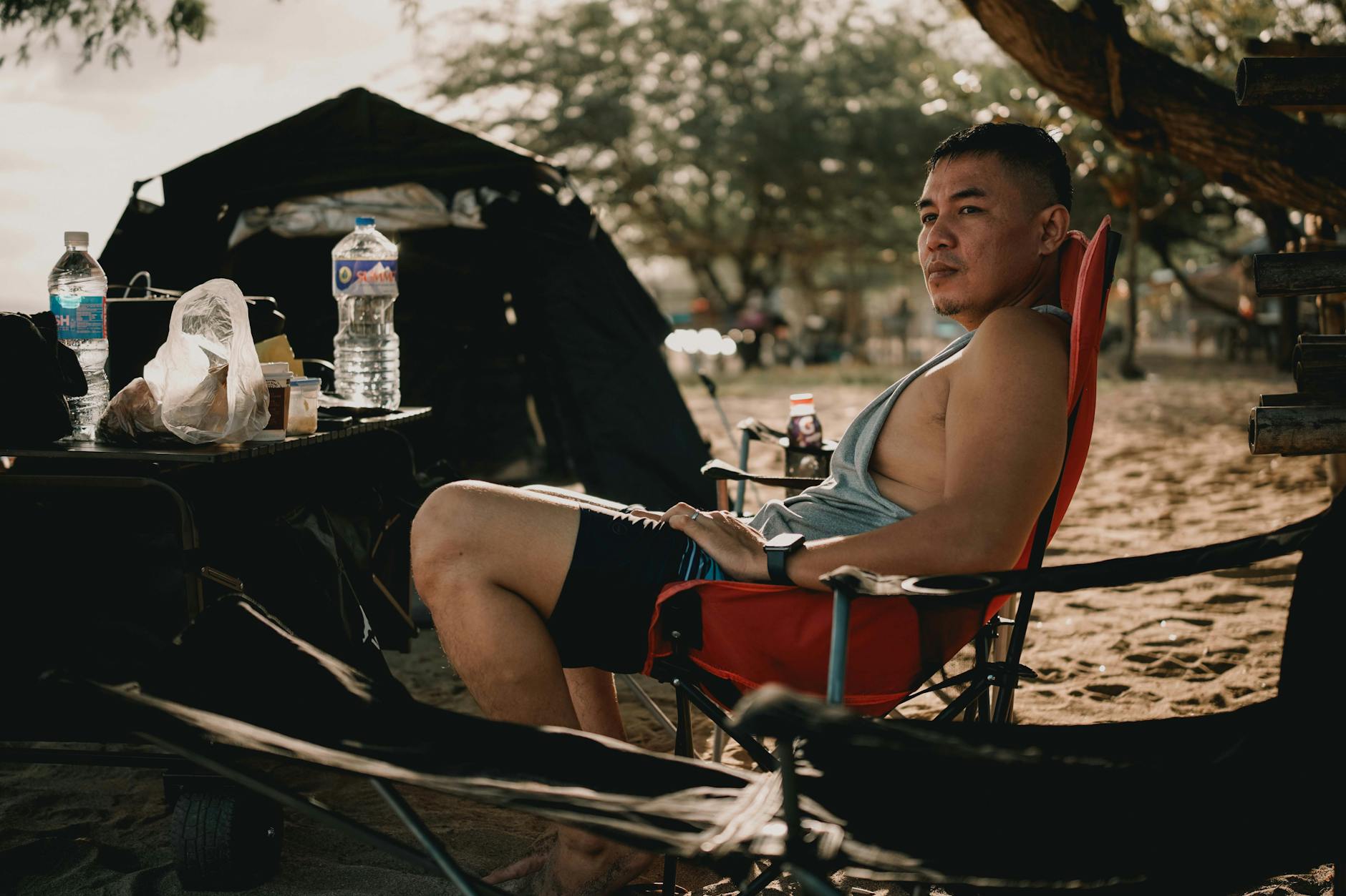 Man relaxing on a beach while camping under a tree with supplies nearby.