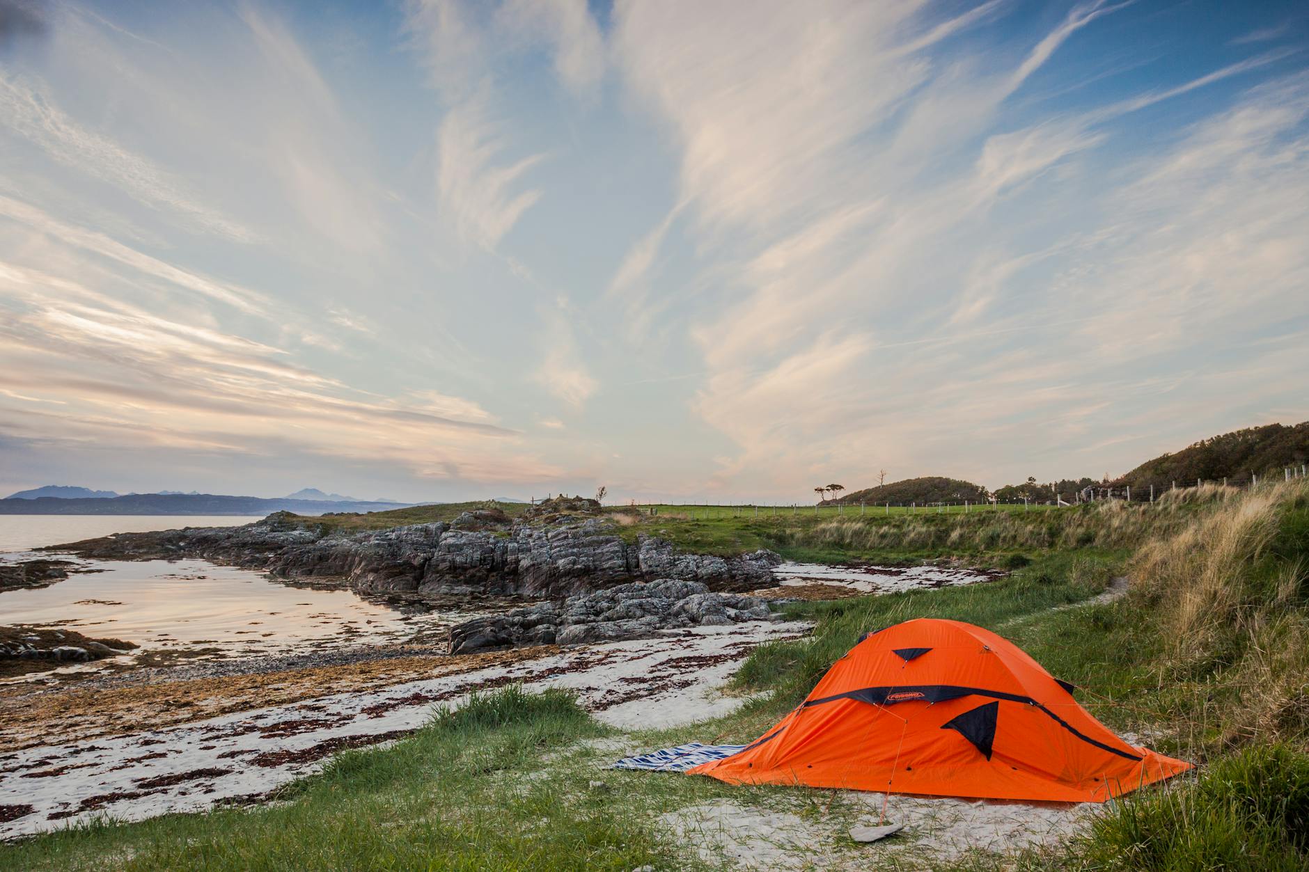 Orange tent set by the serene coast in Highland, Scotland during calm summer evening.