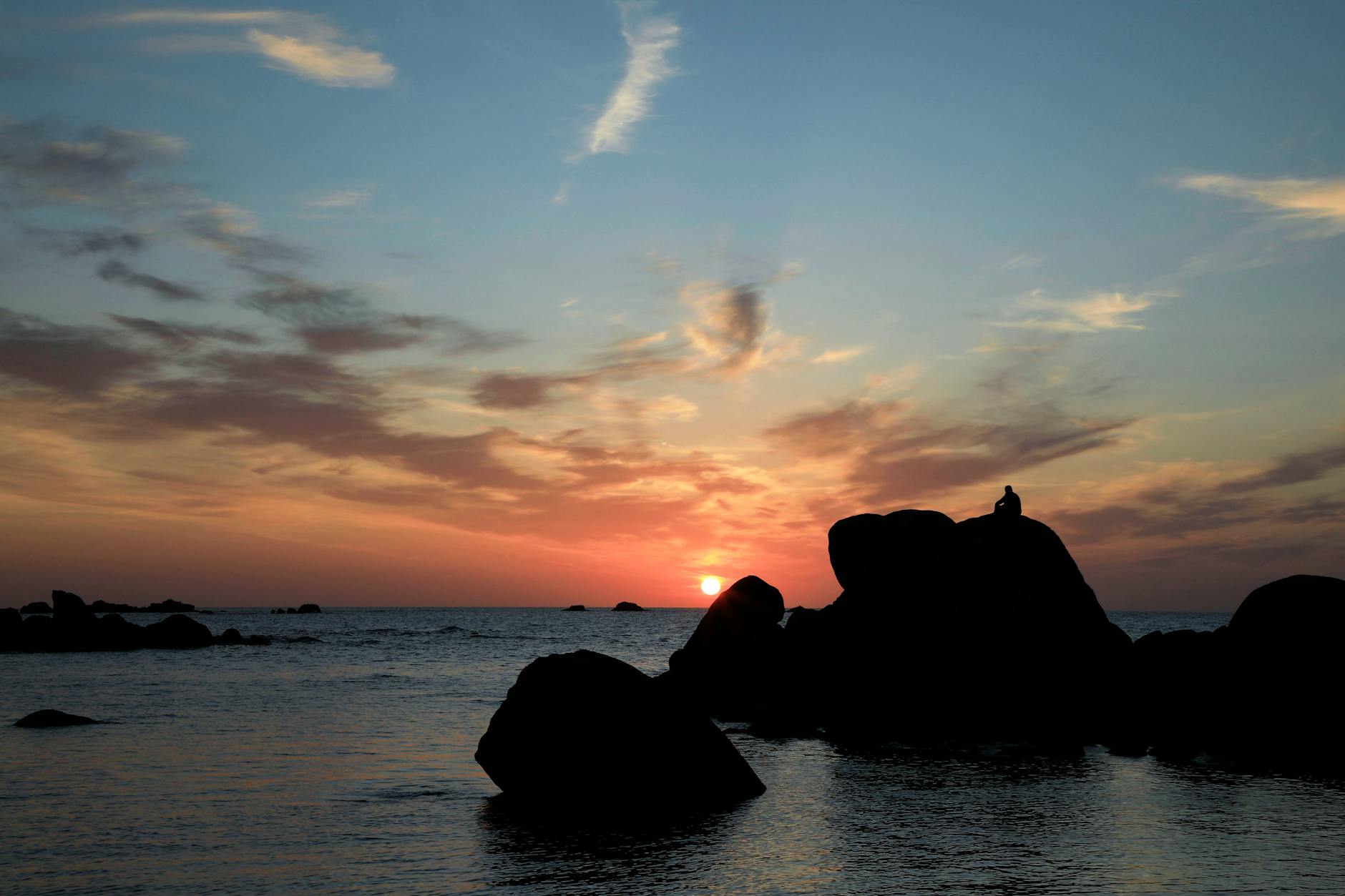 A serene sunset view over the sea with a solitary silhouette on rocks in Bretagne, France.
