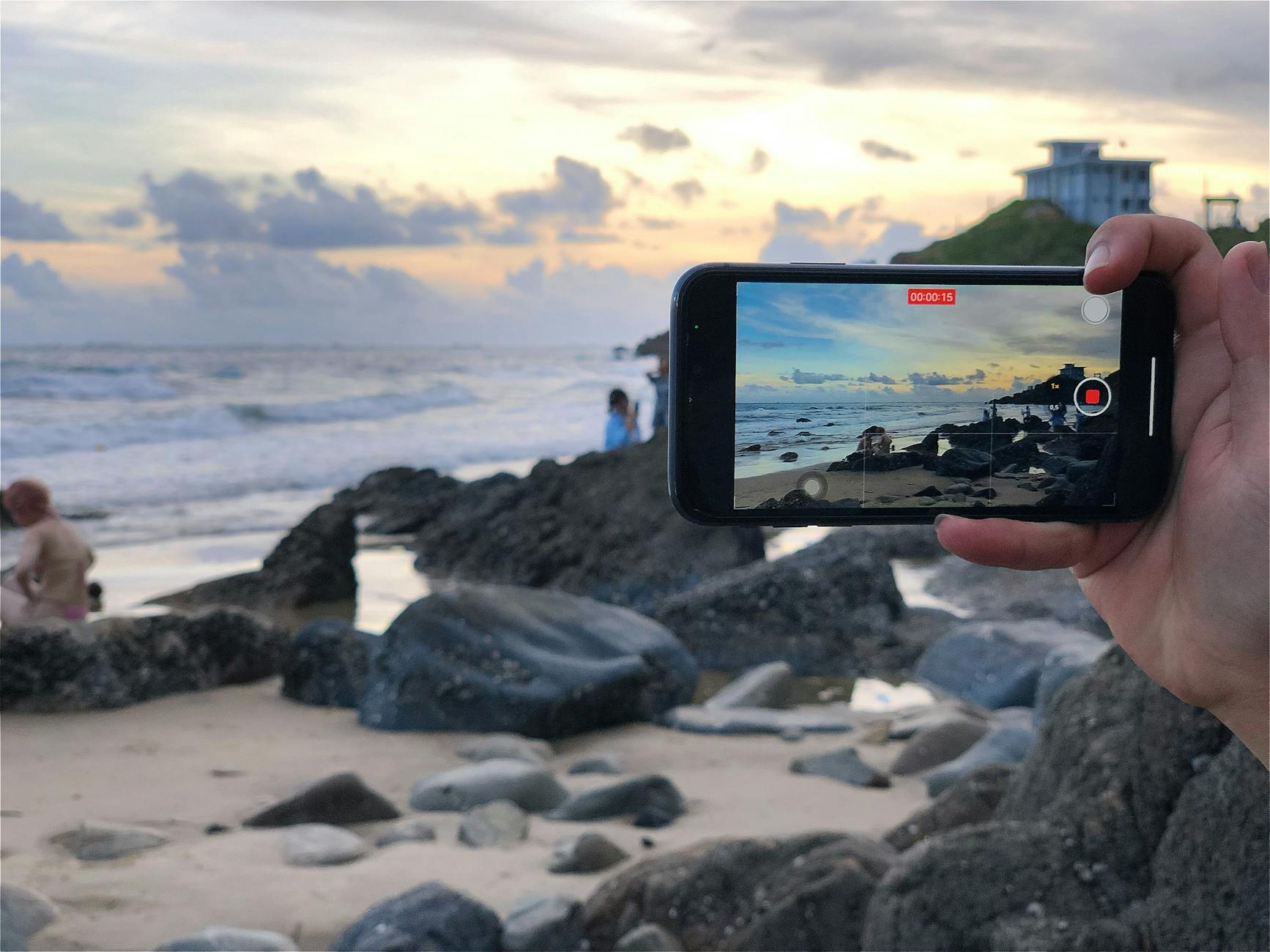 A person records a beautiful beach scene on a smartphone at sunset.