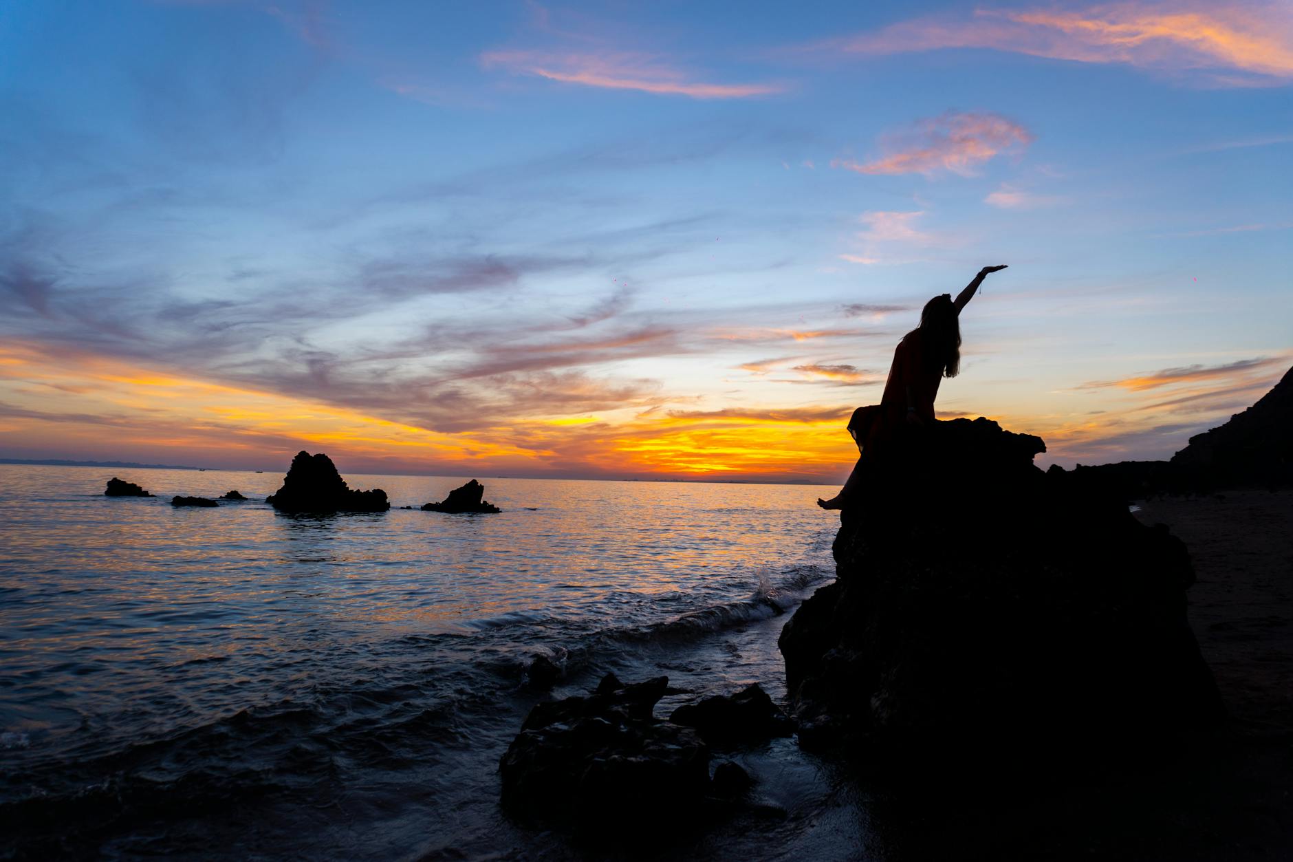 Dramatic silhouette of a person sitting on a rock, waves gently hitting the shore at sunset.