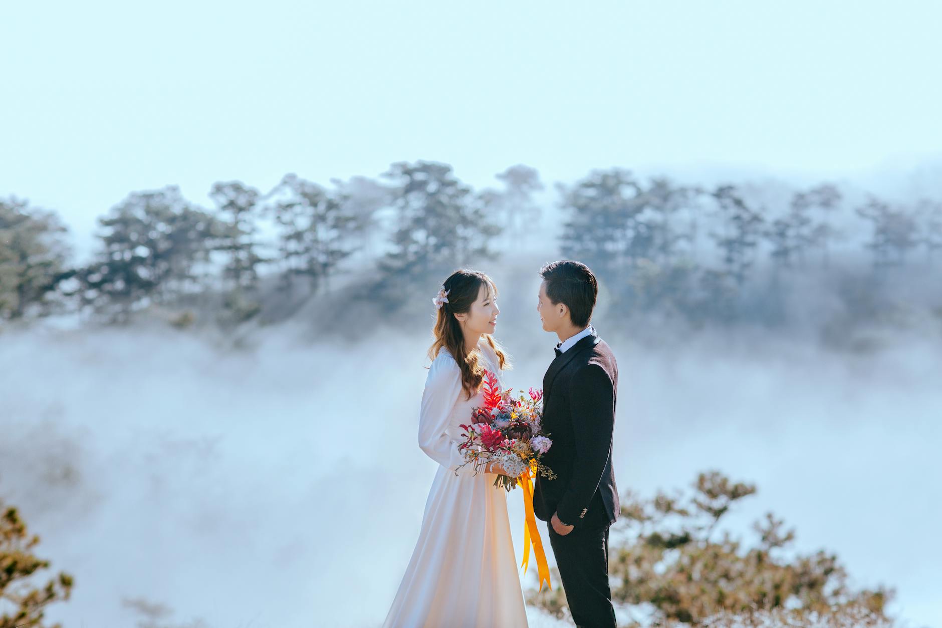 A beautiful Asian couple in wedding attire poses romantically with a scenic foggy forest backdrop.
