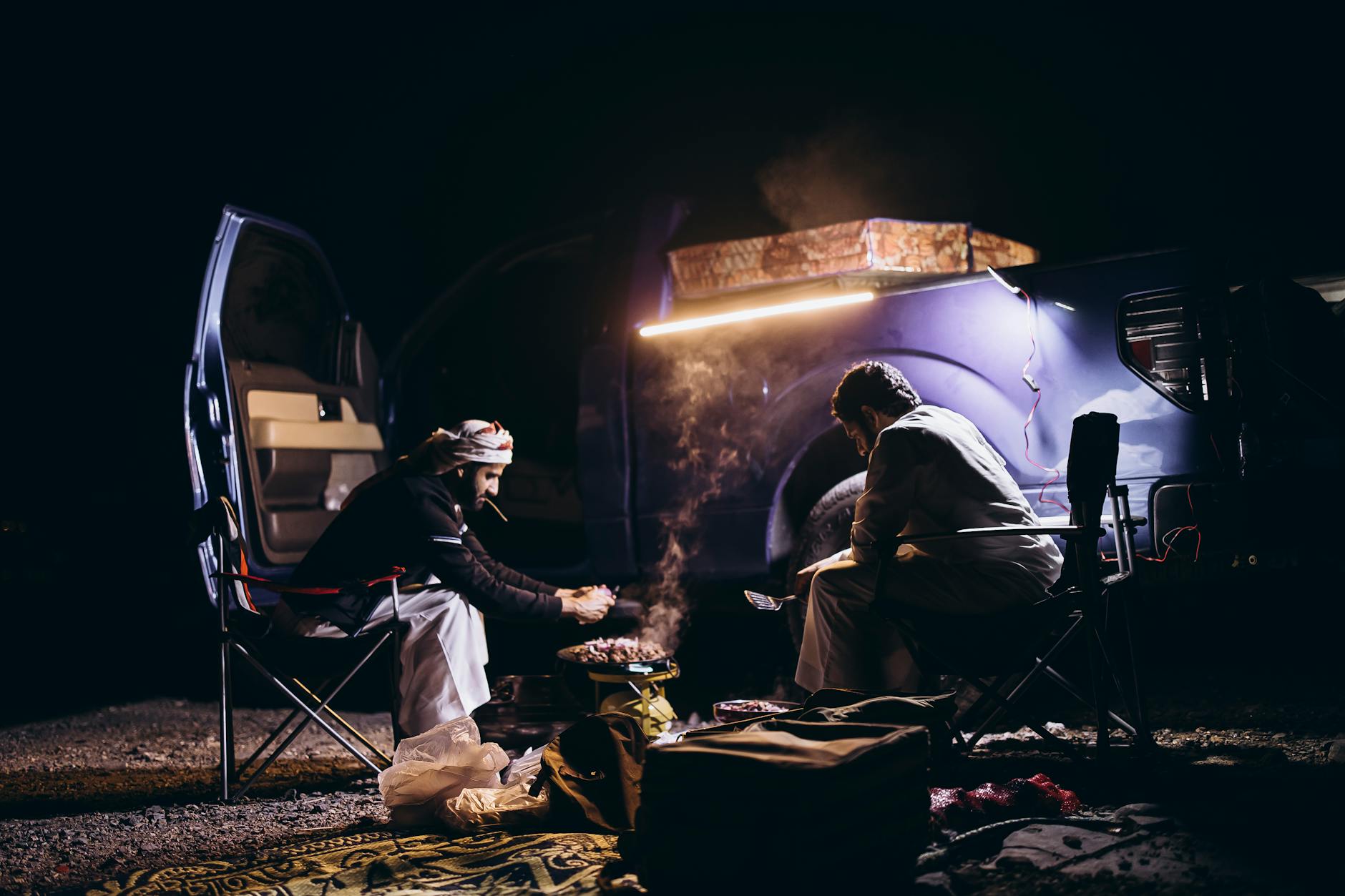 Two men enjoy a cozy night camping and cooking beside a vehicle in Oman's serene wilderness.
