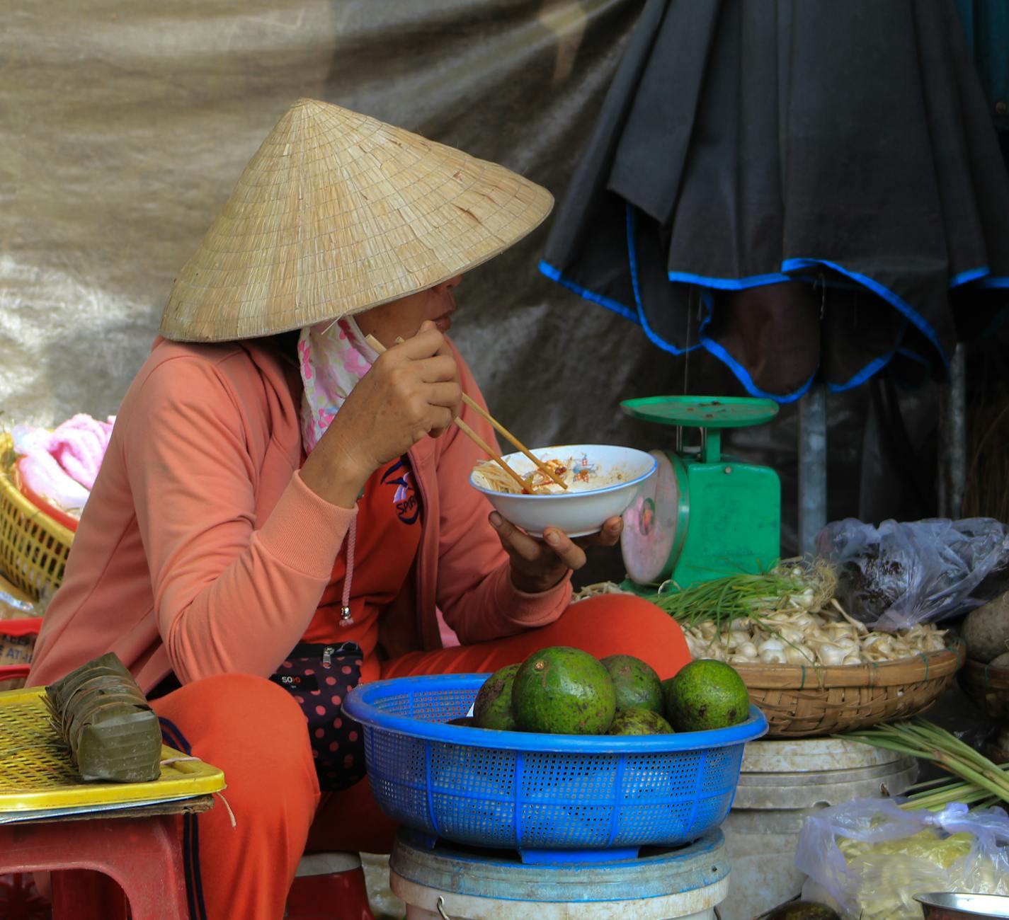 A woman in a Vietnamese street market enjoying a meal while wearing a traditional hat.