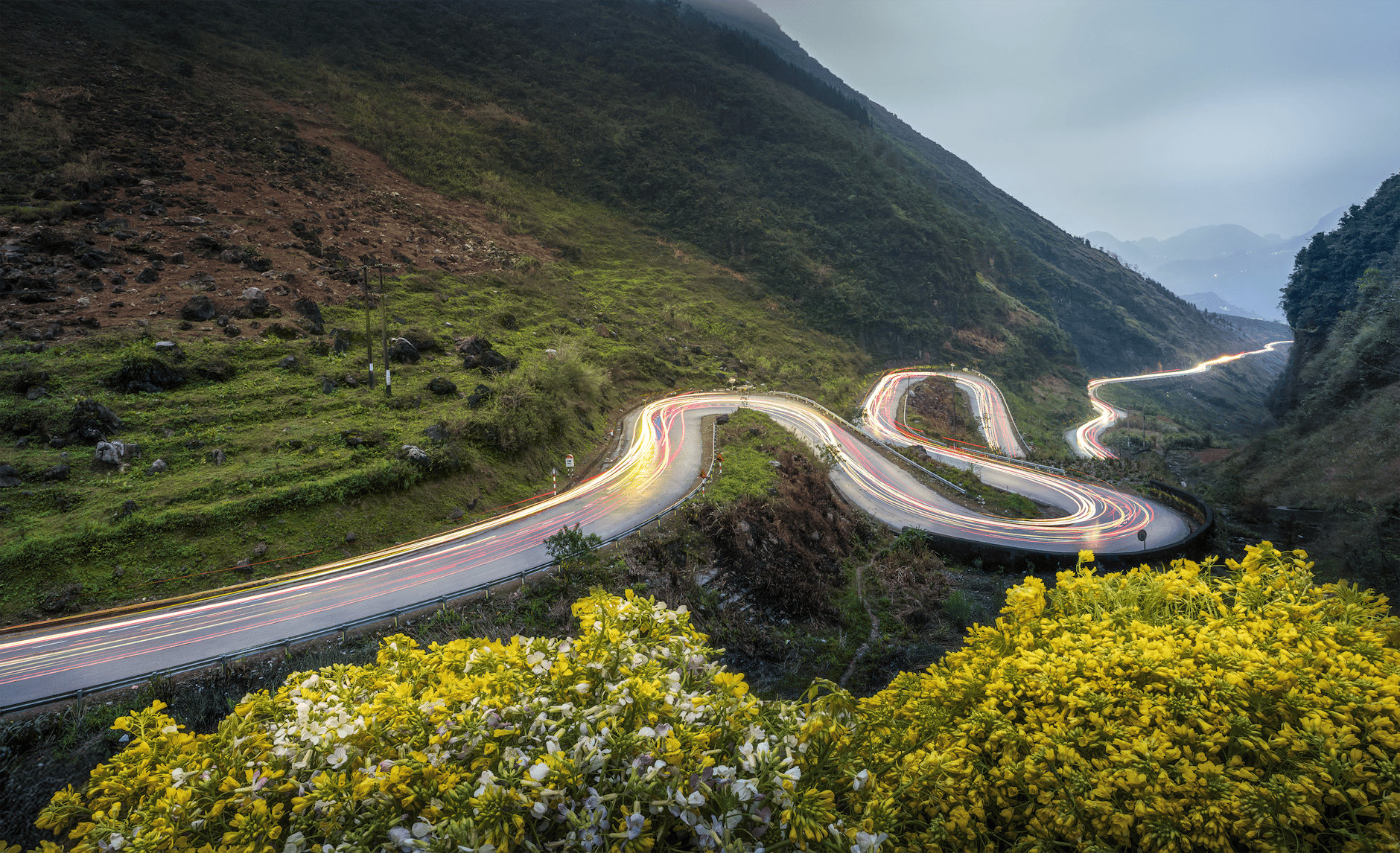 Long-exposure of a winding mountain road with light trails and lush greenery at dusk.