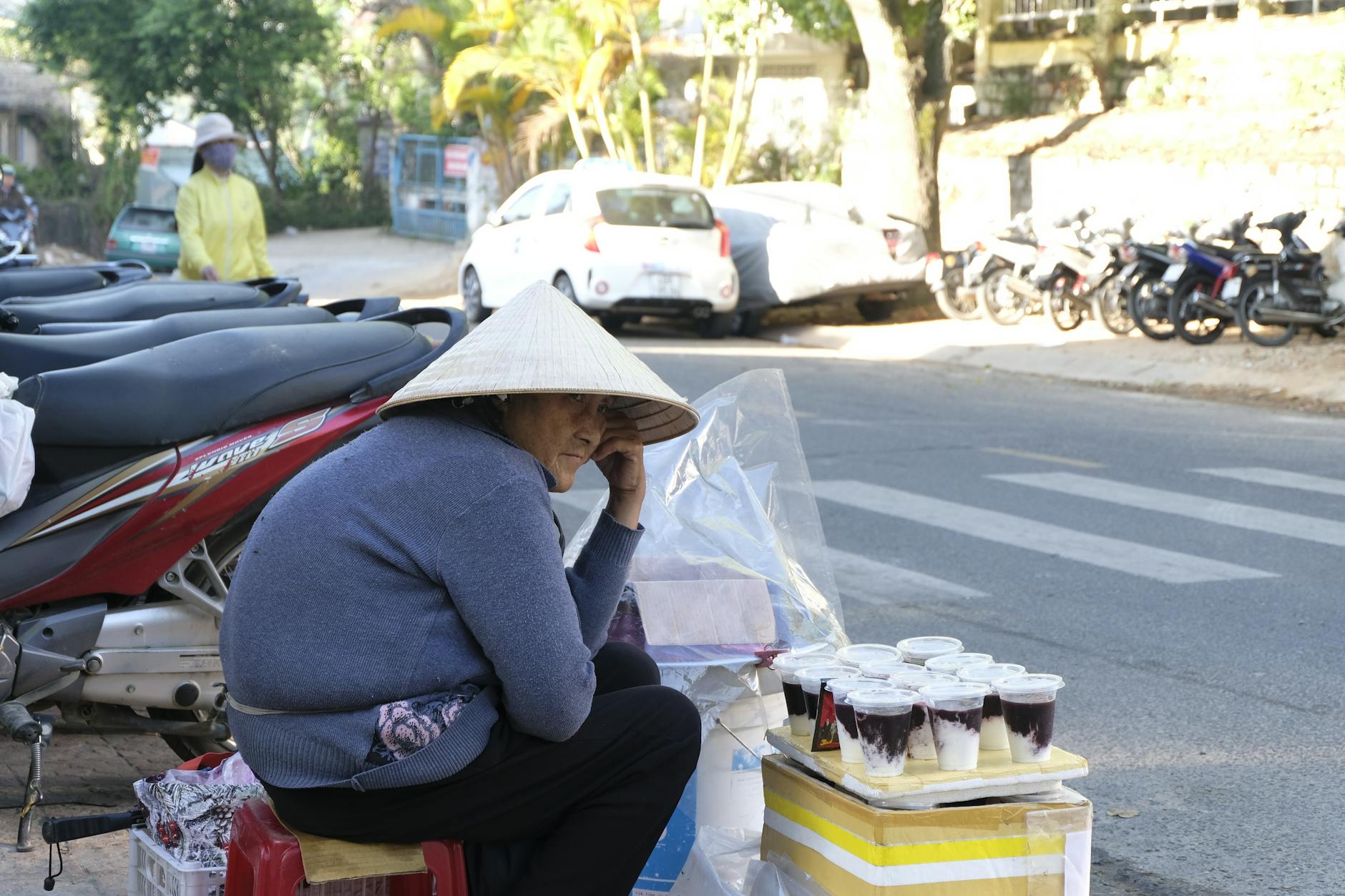 A street vendor with a conical hat sits by the roadside with drinks, showcasing urban street life.