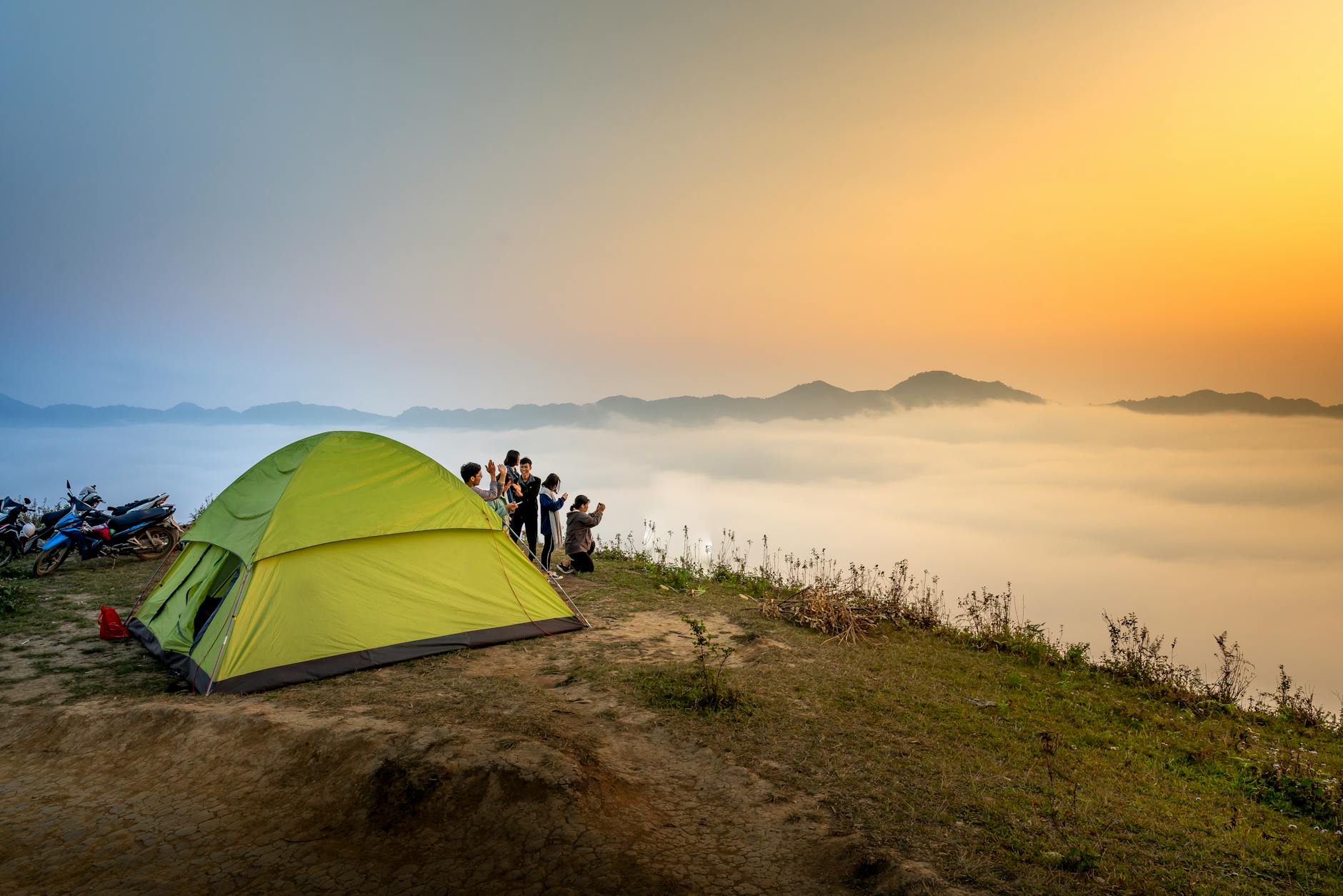 Camping scene with a tent and group at sunrise, overlooking misty mountains.