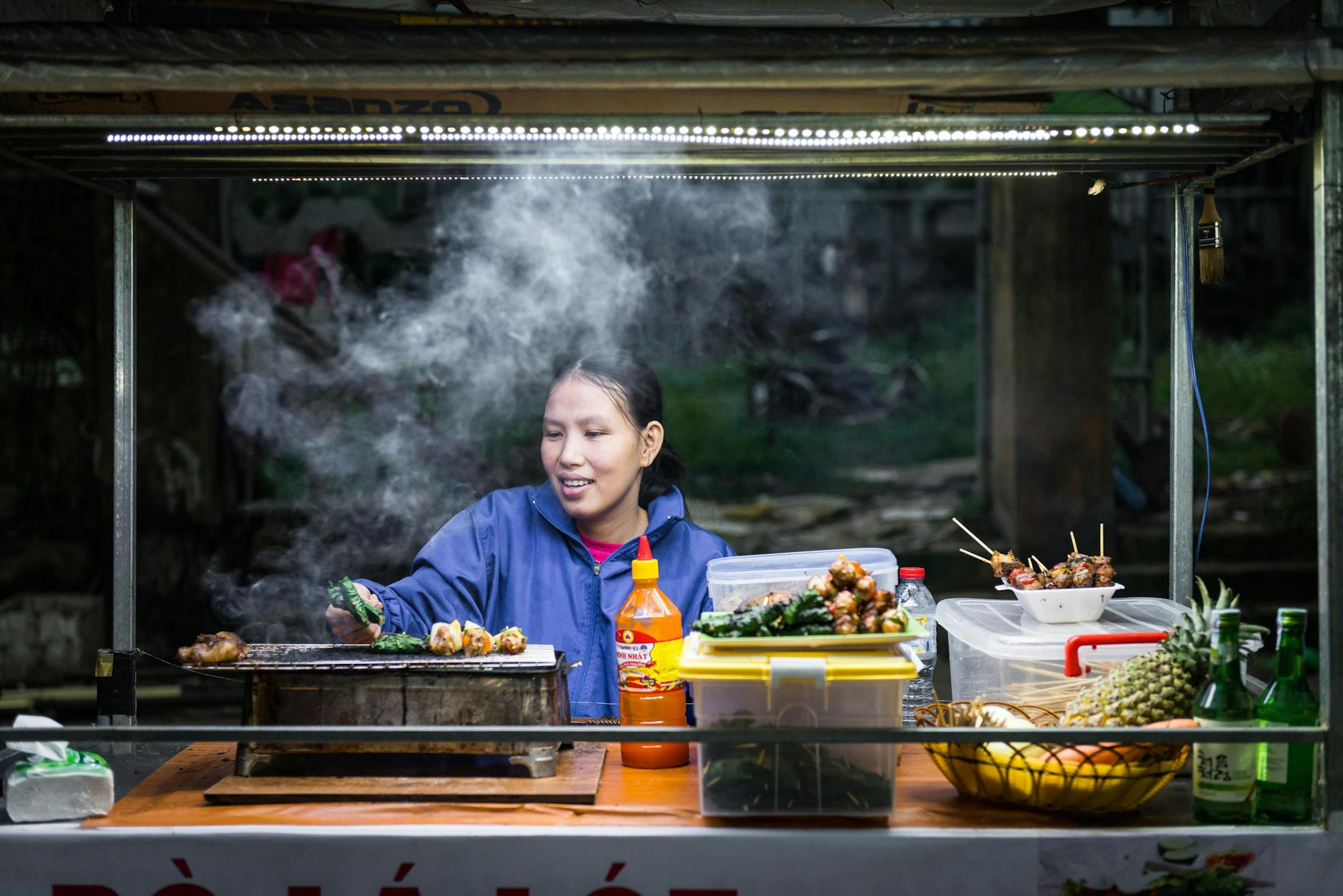 A street food vendor grills skewers amidst steam at a vibrant Hoi An market.