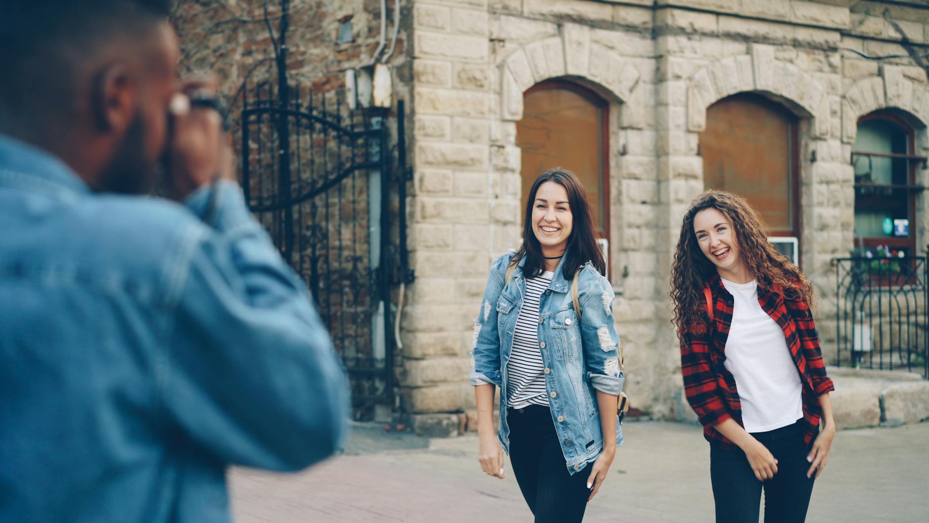 Two young women laughing while a friend photographs them on a city street.