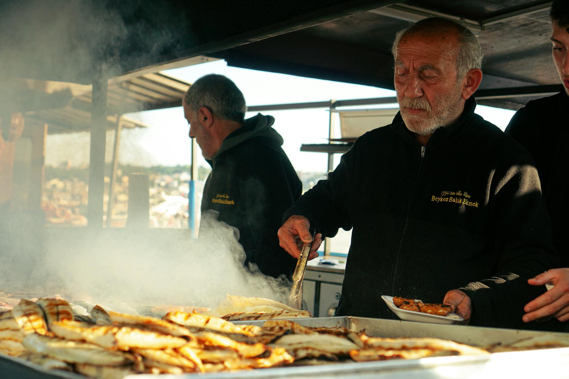 Elderly man grilling fish at an outdoor market, creating a smoky aroma.