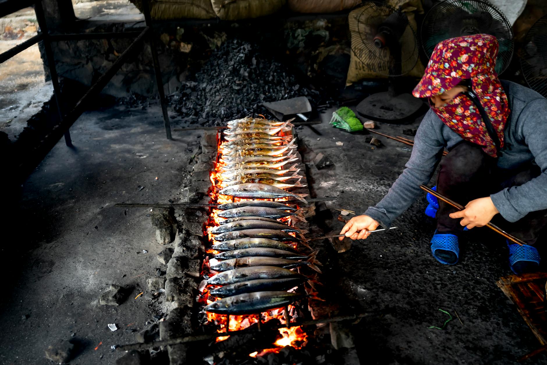 A woman grilling fish over an open charcoal fire, showcasing traditional cooking methods.