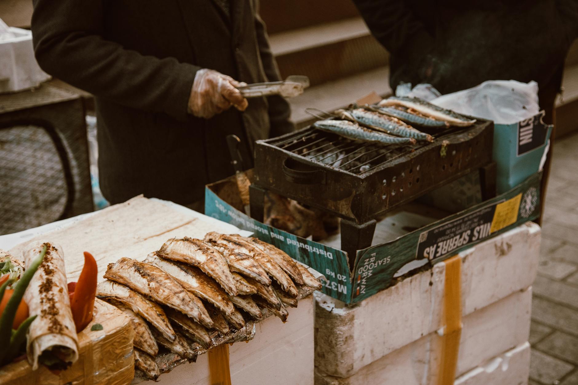 A street vendor grills fresh fish on a portable grill surrounded by fish and vegetables.
