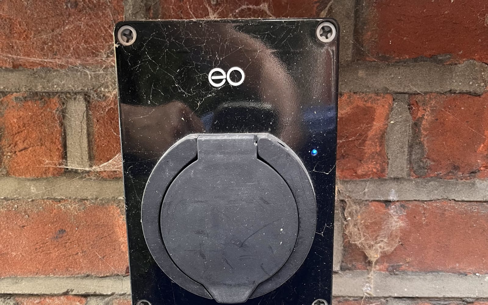 An older square EO Mini Pro home charger mounted on a brick wall with its blue status light showing.