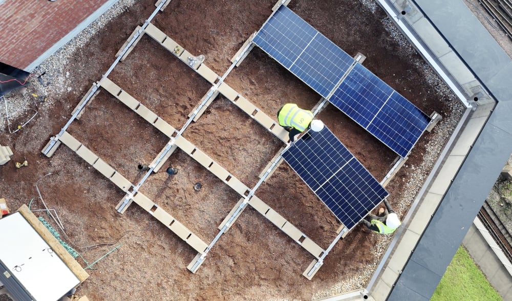 Overhead drone view of a flat roof at Brooke House in Acton, with the aluminium mounting frame laid out and the first solar panels going down, two engineers working on the array.