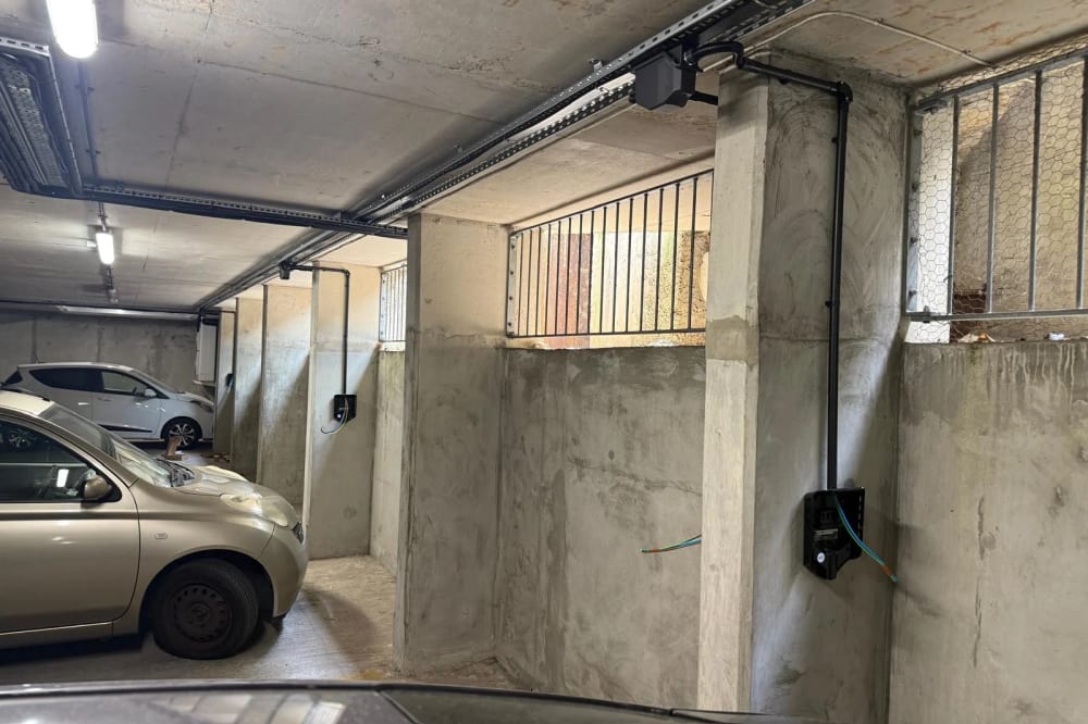 Overhead busbar trunking running the length of an underground car park at Portland Court, with charger units mounted on the concrete bays below and cars parked in between.