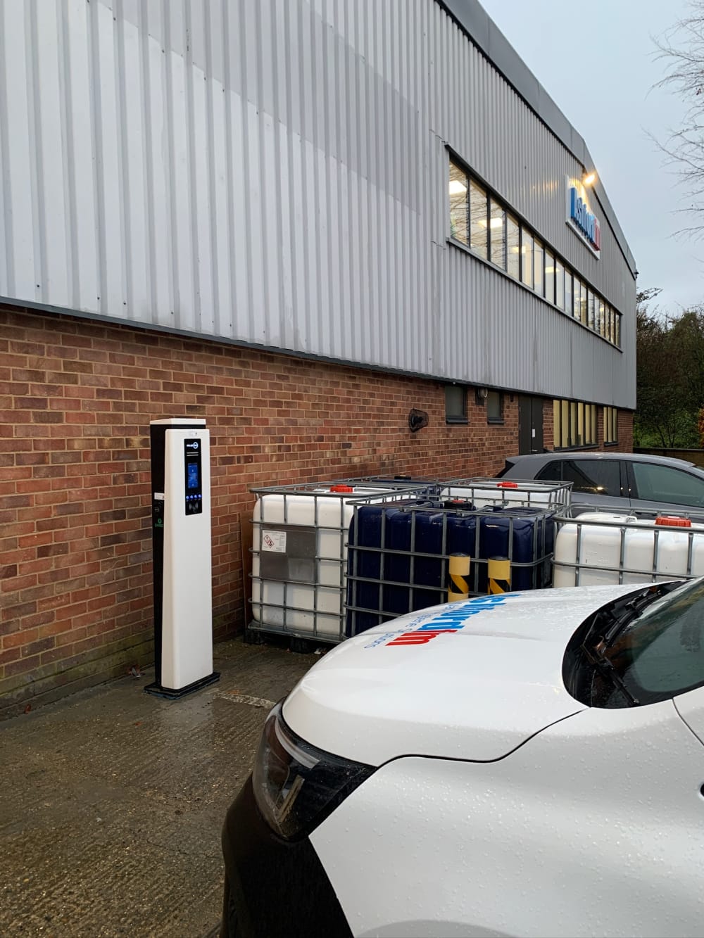 A pedestal EV charger installed outside an industrial unit beside fleet vehicles and storage tanks in the UK.