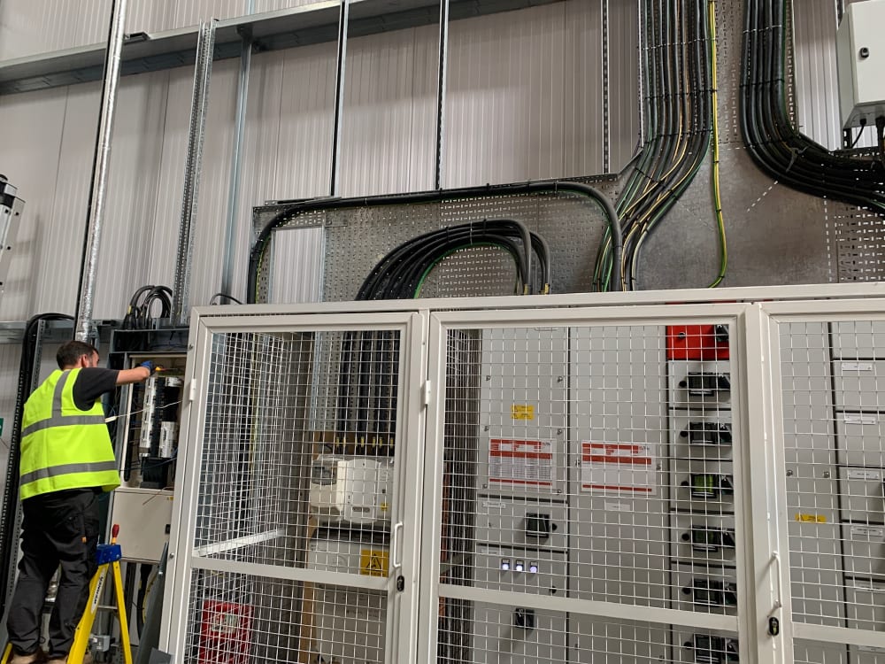 An electrician installing cable containment and distribution switchgear for a commercial EV charging power upgrade in the UK.