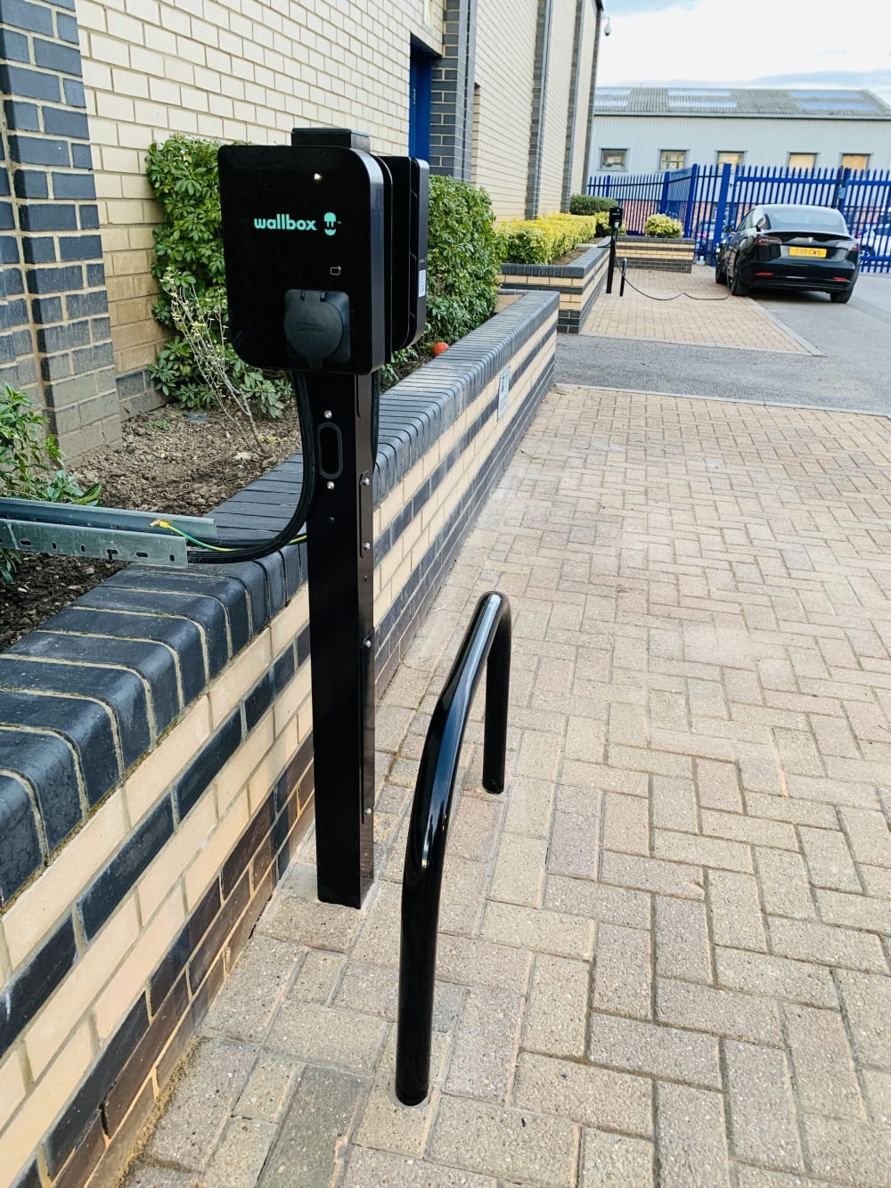 A Wallbox EV charger on a post with wheel guard installed in a business car park beside parked cars in the UK.