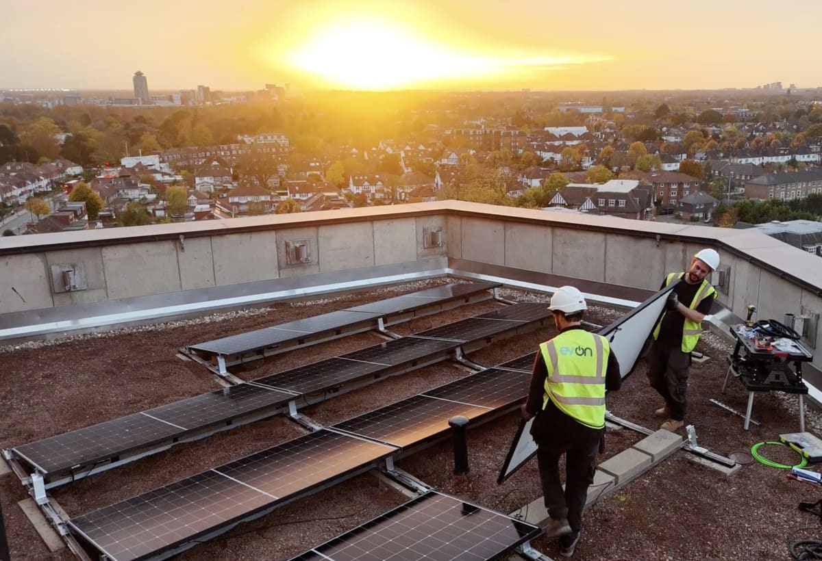 EV-ON engineers in branded hi-vis carrying solar panels at sunset on the 13th floor roof of Brooke House, Acton, during the commissioning stage.