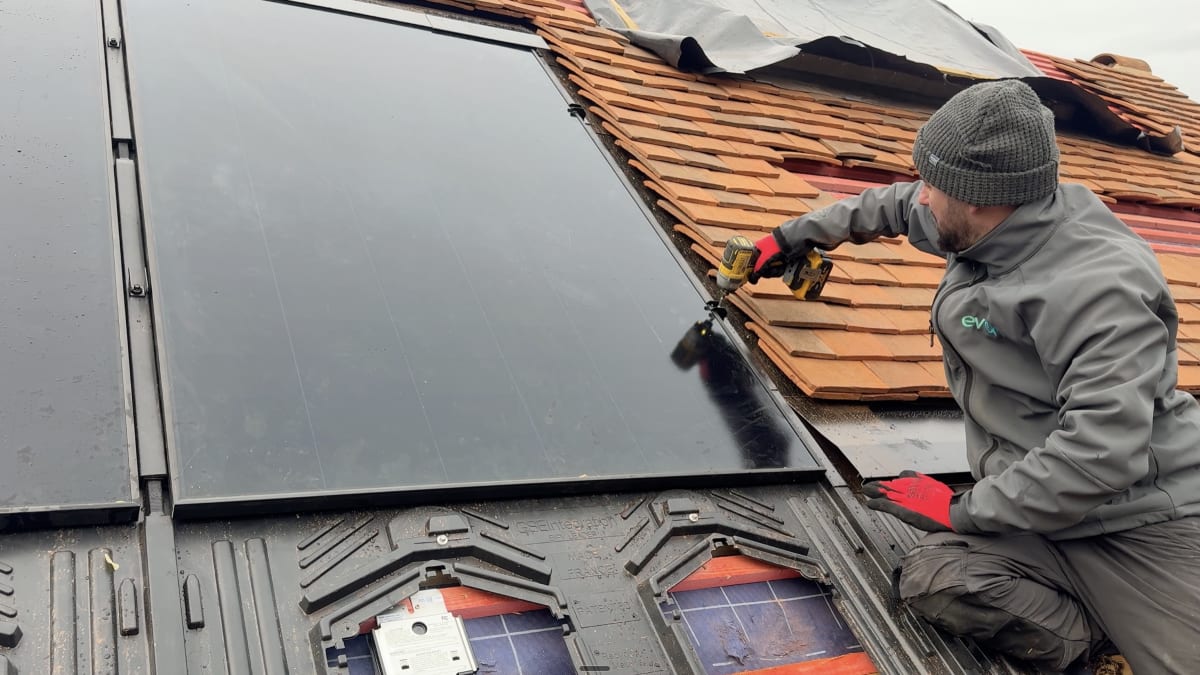 EV-ON engineer installing an Aiko all-black solar panel on a GSE in-roof tray at a detached cottage in Broxted, Essex, with tree shading visible in the background.