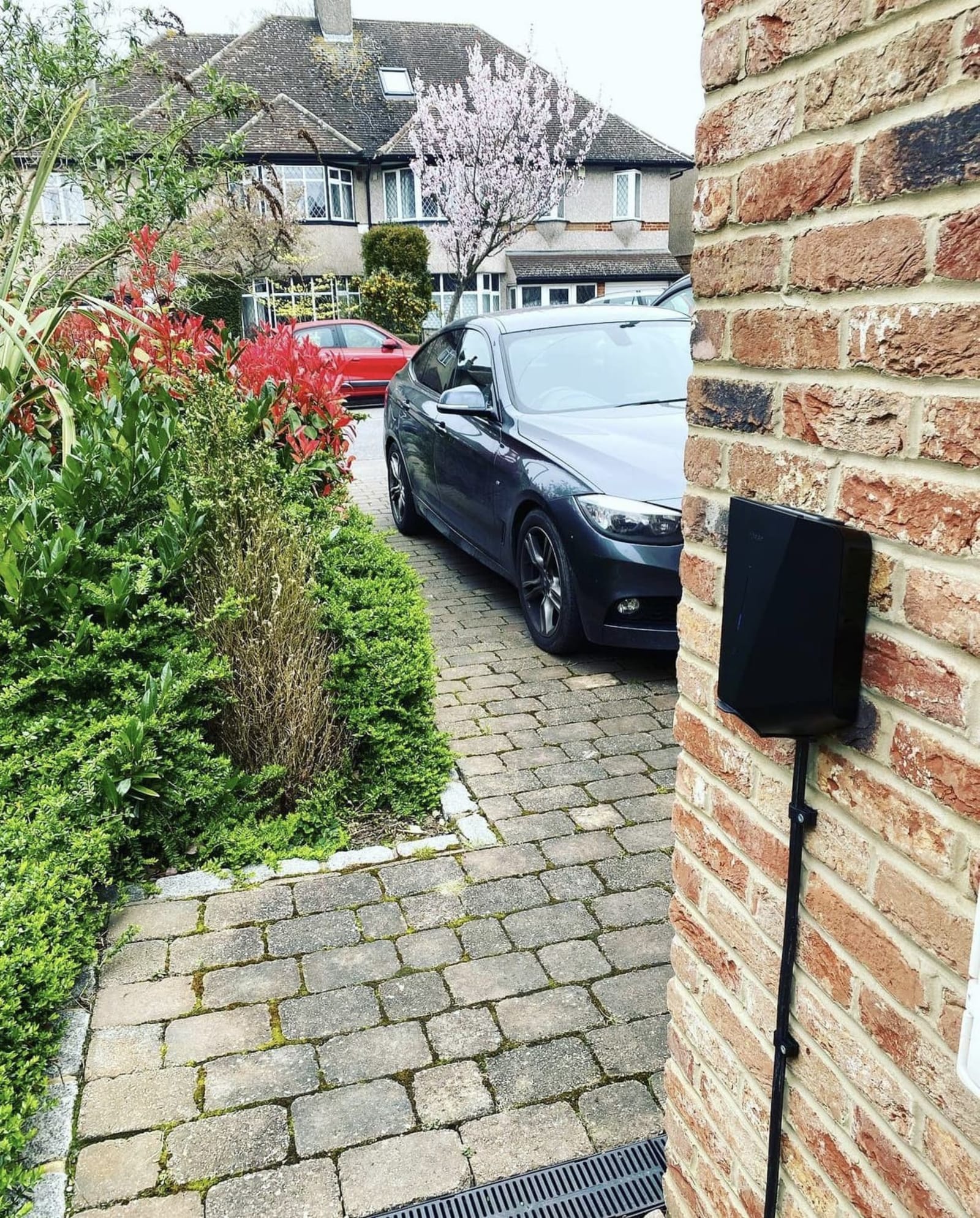 A black Easee One home EV charger fitted to a brick wall beside a car on a residential driveway in the UK.