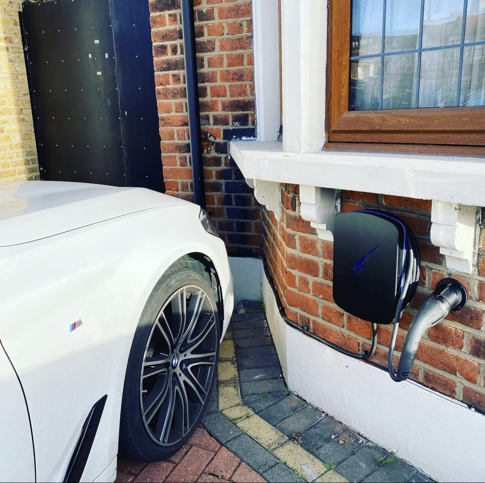 A black Hypervolt EV charger fitted under a window on a brick house wall next to a parked car in the UK.