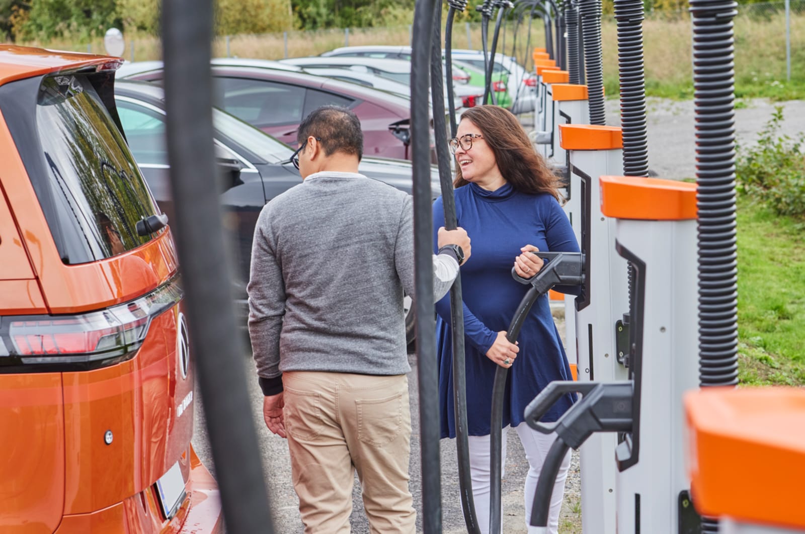 Two people at a Kempower commercial EV charging station with satellite chargers and orange electric vehicles.