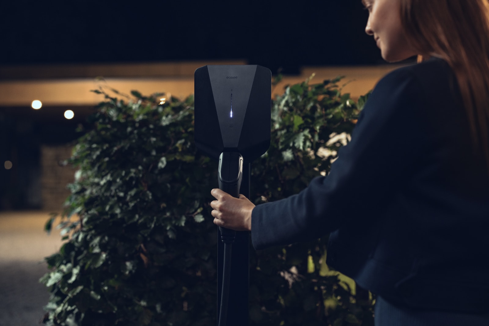 A woman takes the connector from a sleek black Easee pedestal EV charger outside a softly lit hotel at dusk