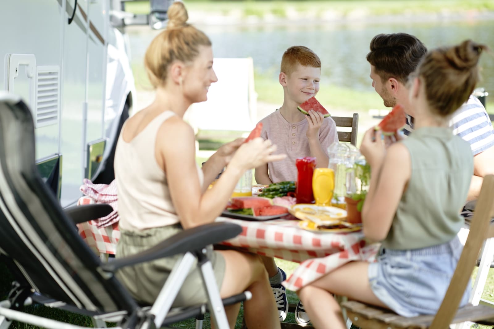 A family eating at a table beside their motorhome next to a lake on a summer camping holiday.