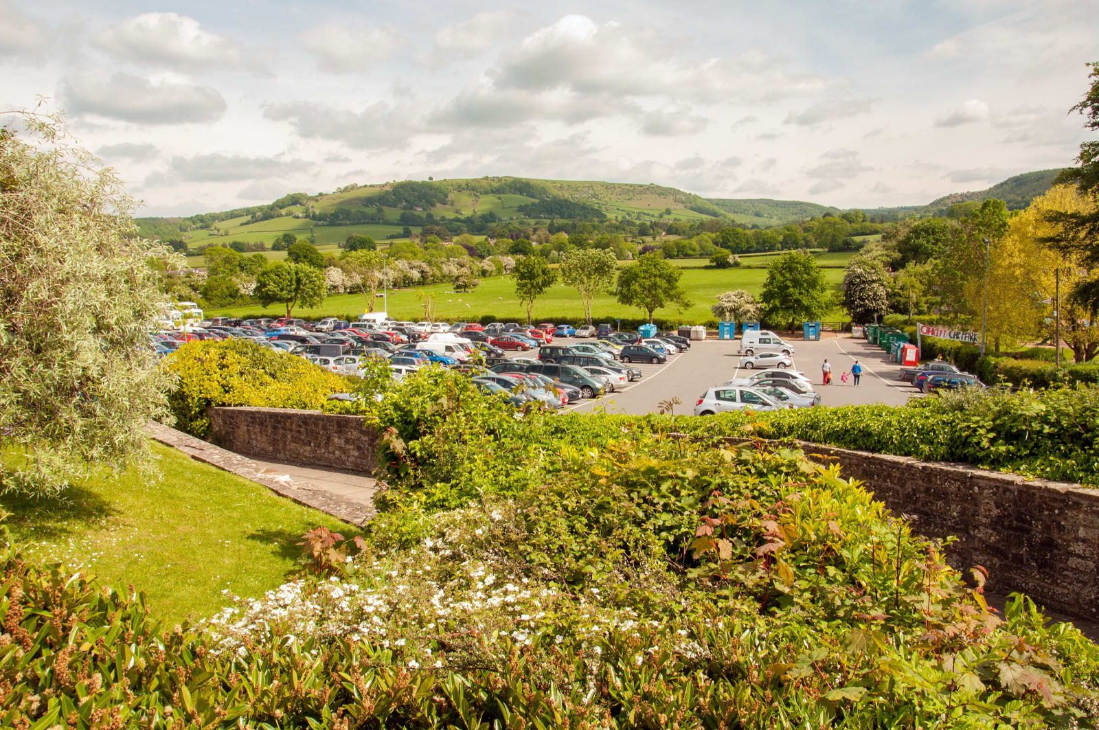 A busy rural visitor car park full of cars with green hills behind on a sunny day.