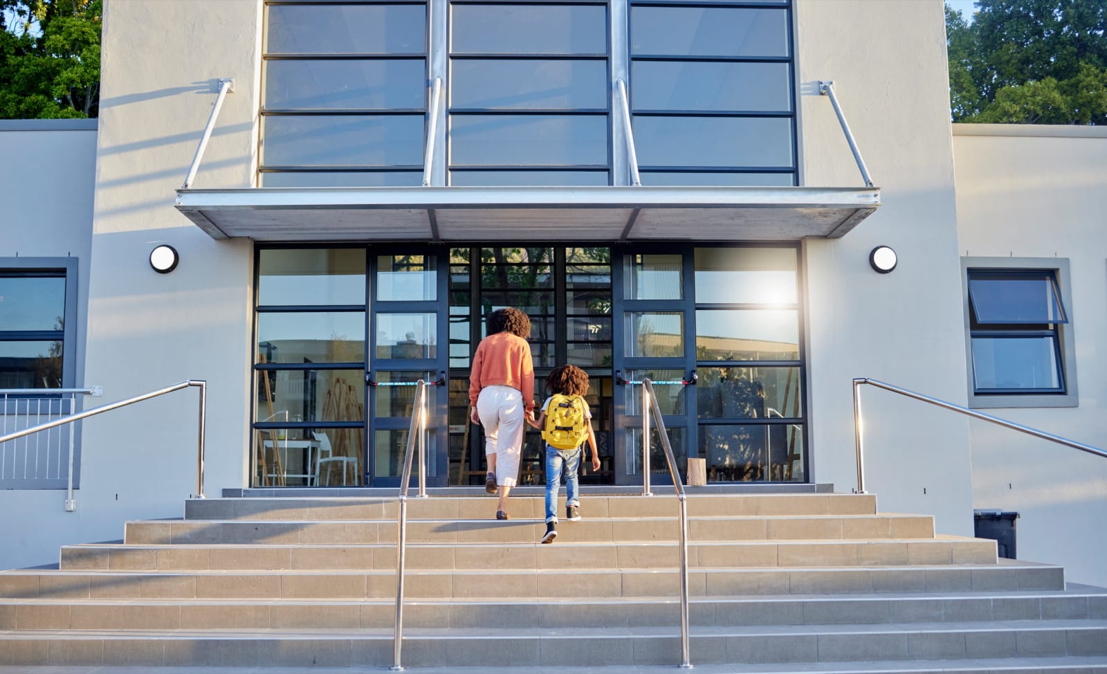 A parent walking a child with a yellow backpack up the steps into a modern school entrance.
