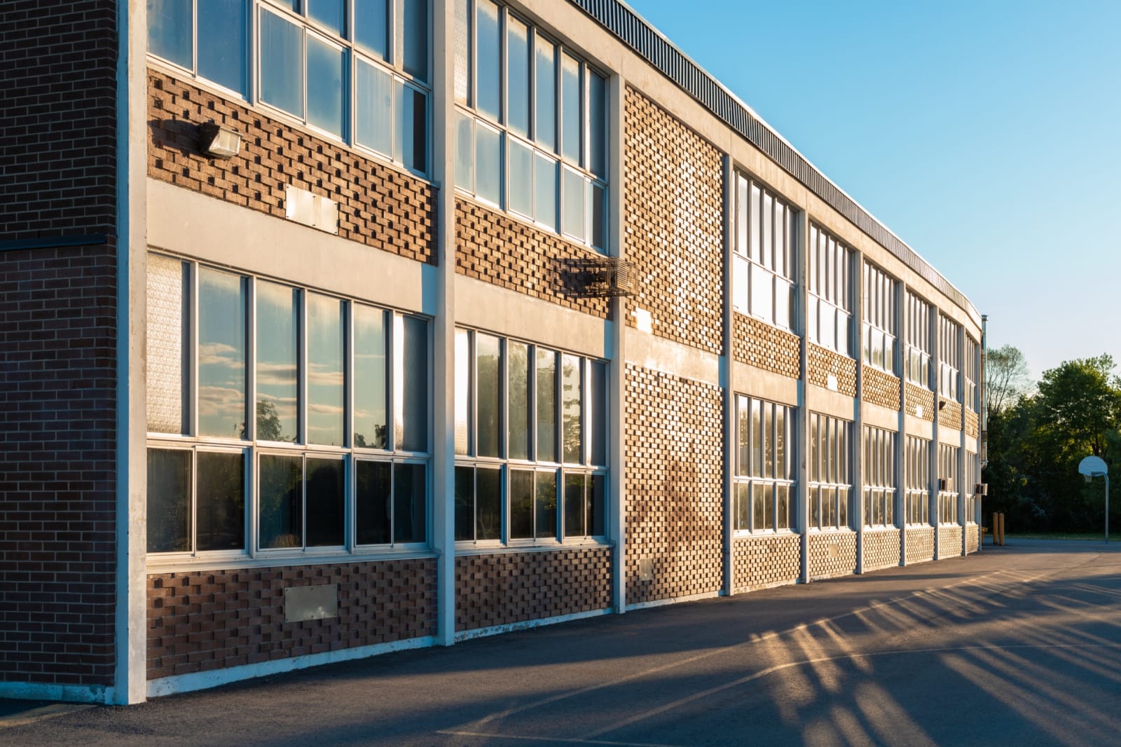 A brick school building and tarmac schoolyard with a basketball hoop in golden evening light.