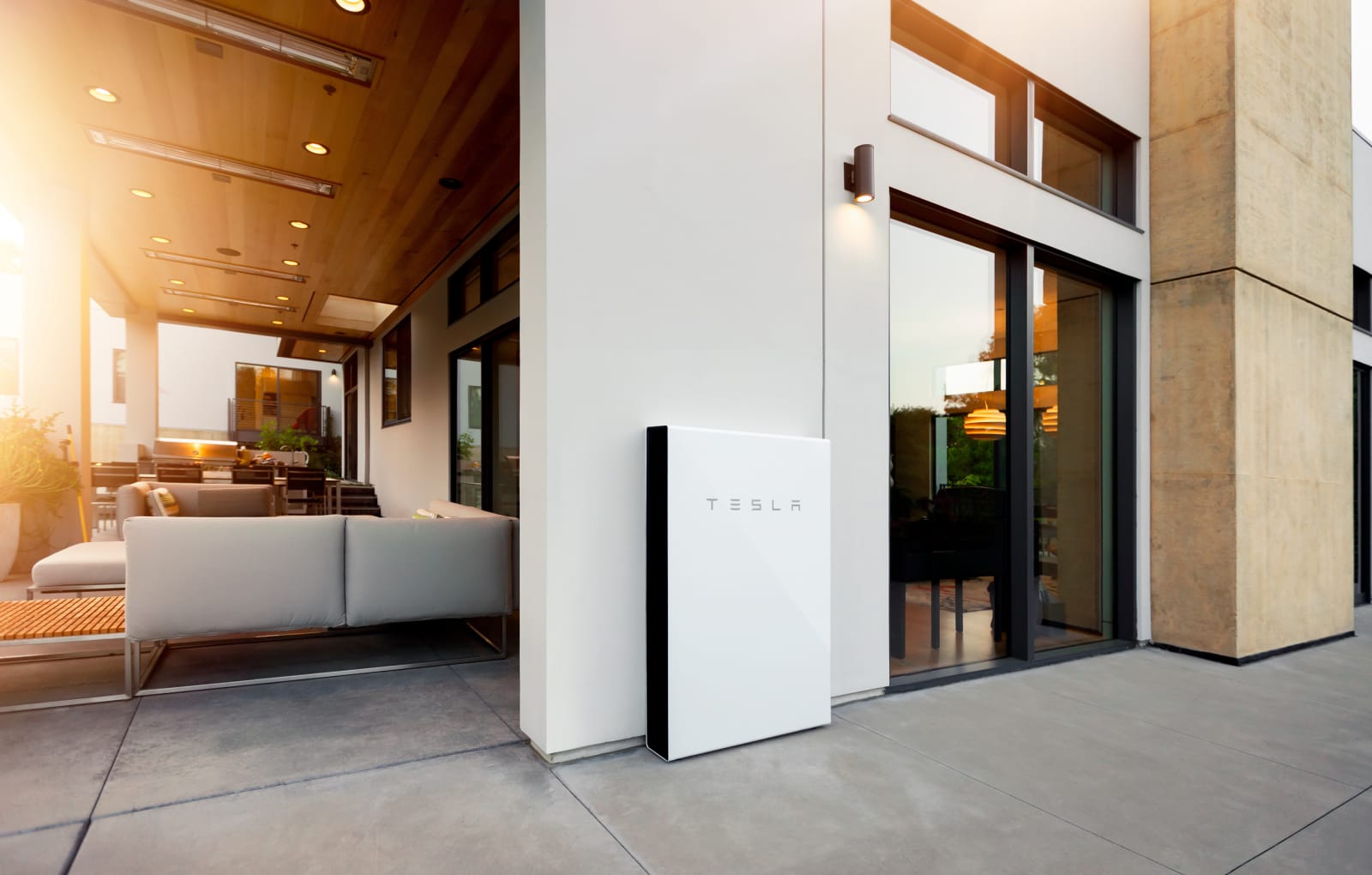 Tesla Powerwall battery storage unit installed on an exterior wall beside glass sliding doors into an open plan living space.