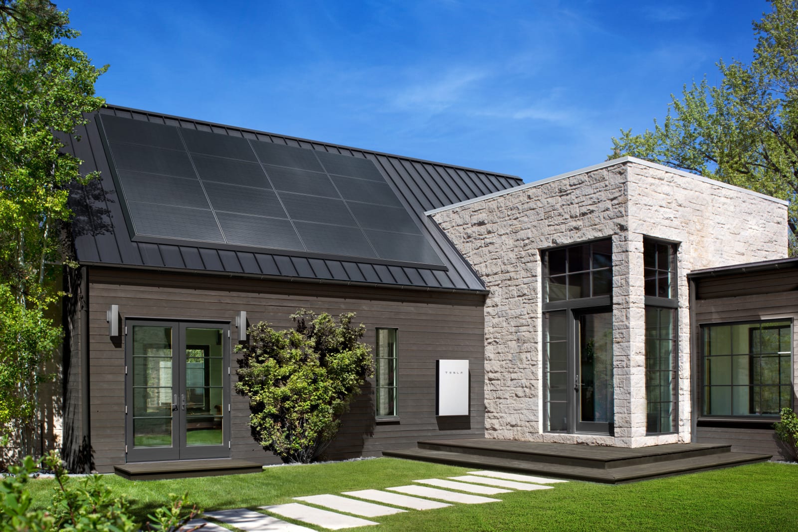 Tesla solar panels fitted to the black roof of a stone clad modern home with a Tesla Powerwall visible outside.