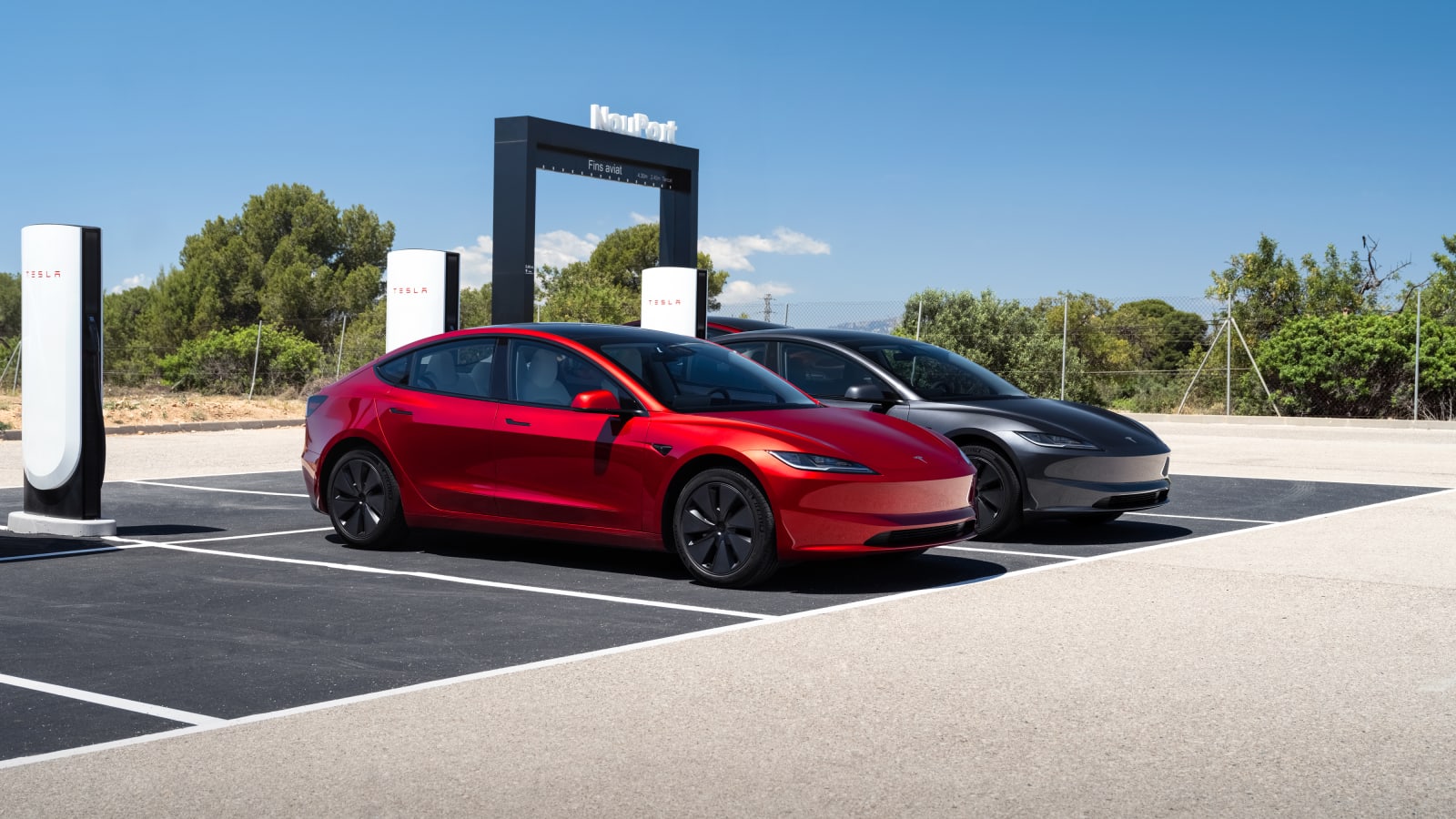 Red and grey Tesla cars parked at a Tesla Supercharger bay with an illuminated sign in the background.
