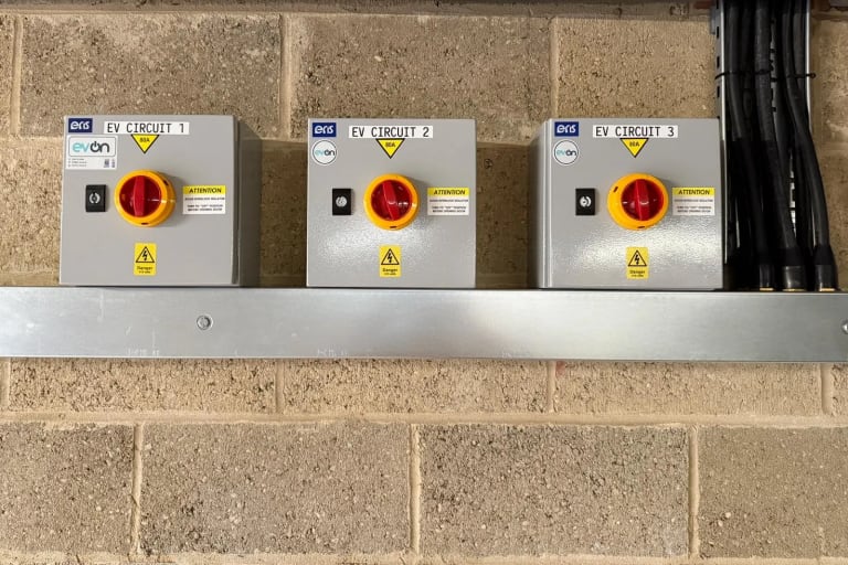 Three grey isolator enclosures labelled EV Circuit 1, 2 and 3, each with a red rotary switch, mounted on a steel rail against a blockwork wall at Portland Court.