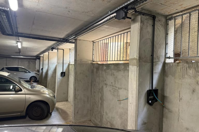 Overhead busbar trunking running the length of an underground car park at Portland Court, with charger units mounted on the concrete bays below and cars parked in between.