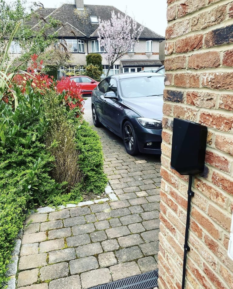 A black Easee One home EV charger fitted to a brick wall beside a car on a residential driveway in the UK.