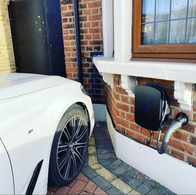A black Hypervolt EV charger fitted under a window on a brick house wall next to a parked car in the UK.