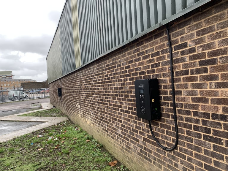 A black wall mounted EV charger fitted to the brick exterior of an industrial unit beside a car park in the UK.