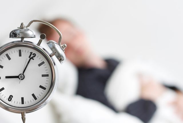 Journée nationale du sommeil du 14 mars 2025. Vue d'un réveil avec en arrière plan une personne allongée qui sort du sommeil.