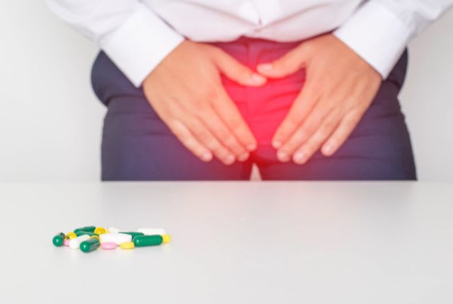 Priapisme. Un homme tient son bas ventre de douleur, celui-ci est teinté de rouge pour marquer l'intensité du trouble. Devant lui, des médicaments sont étalés sur une table.