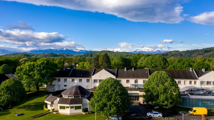 Aerial view of a healthcare facility surrounded by green trees, with mountains in the background under a partly cloudy blue sky.