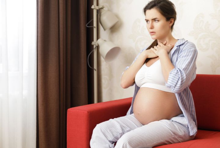 Mal de gorge chez la femme enceinte : image d'une femme enceinte assise sur un canapé se tenant la gorge, car souffrant d'un mal de gorge