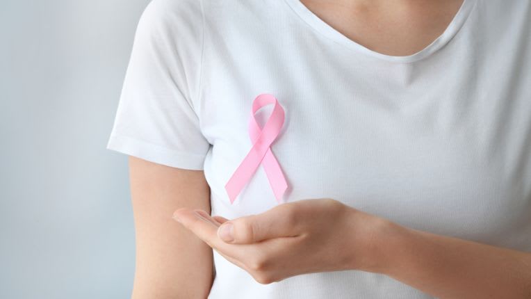 Cancer du sein. Une femme en t-shirt blanc dont on ne voit pas le visage porte un ruban rose sur le sein droit et pose sa main juste en dessous comme pour le soutenir.