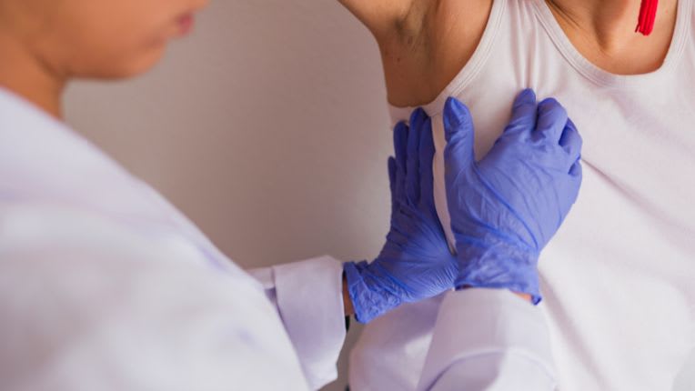 Cancer du sein : combien de vies sauvées grâce au dépistage selon l'Inca ? Gros plan sur les mains d'une soignante, gantées, pratiquant une palpation du sein d'une patiente, au travers de son t-shirt blanc.